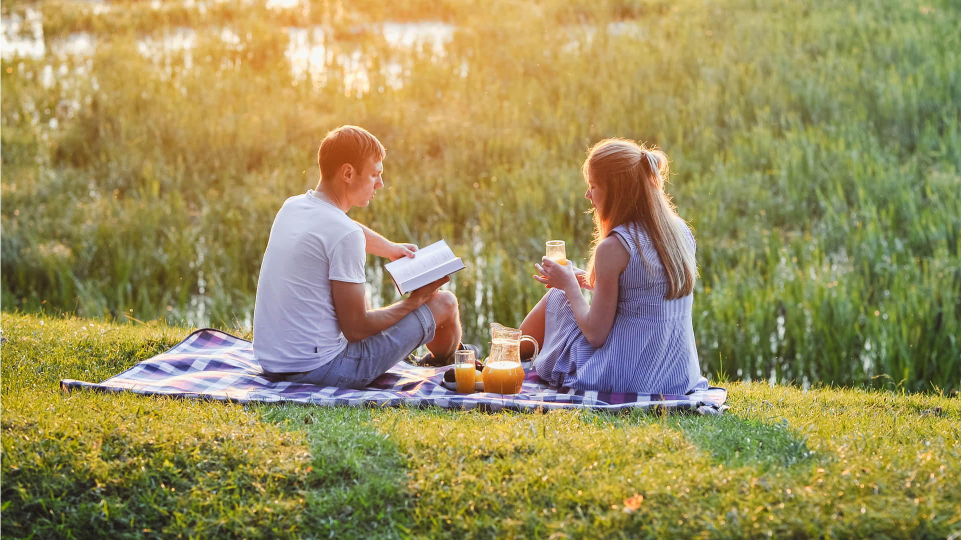 Man in white shirt reading book sitting down in a park with woman next to him drinking glass of orange juice.