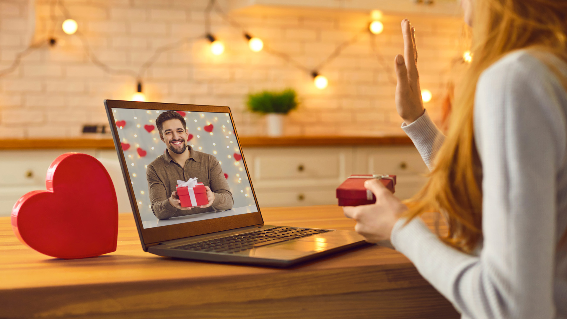 Woman holding a small red box waving at her laptop screen showing a man also holding a red box. A red love-heart on the table