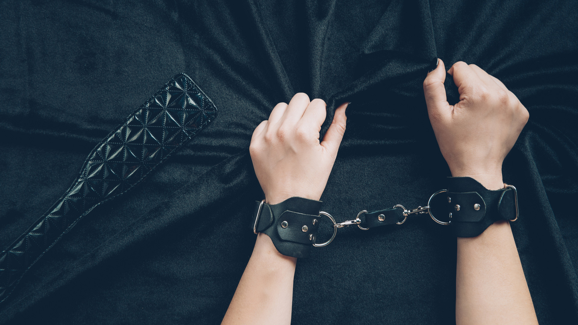 Hands of a woman cuffed together gripping black sheets with a spanking paddle right next to them.